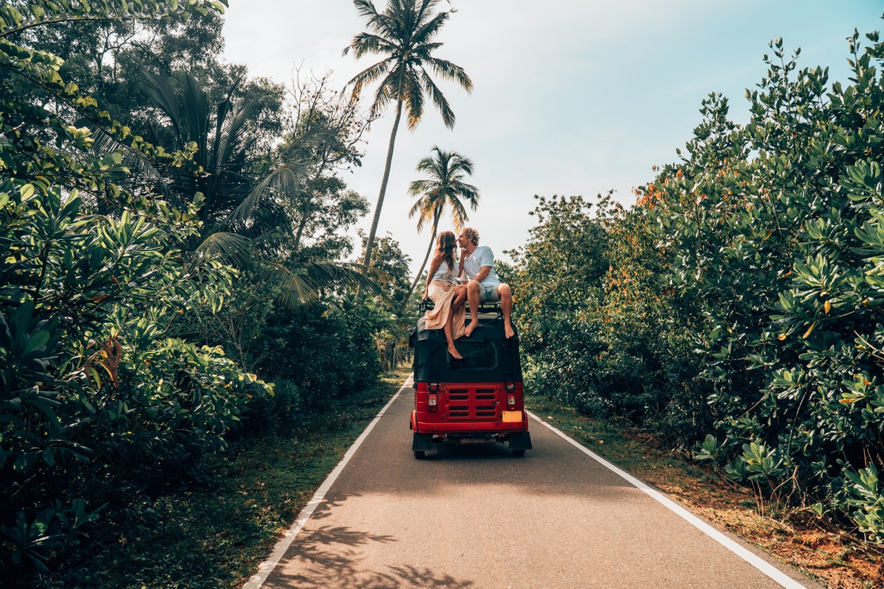 Young adult couple on a tropical summer adventure road trip journey in Sri Lanka. Rented red Tuk Tuk vehicle. Tropical road surrounded by palm trees. People travel exploration concept
