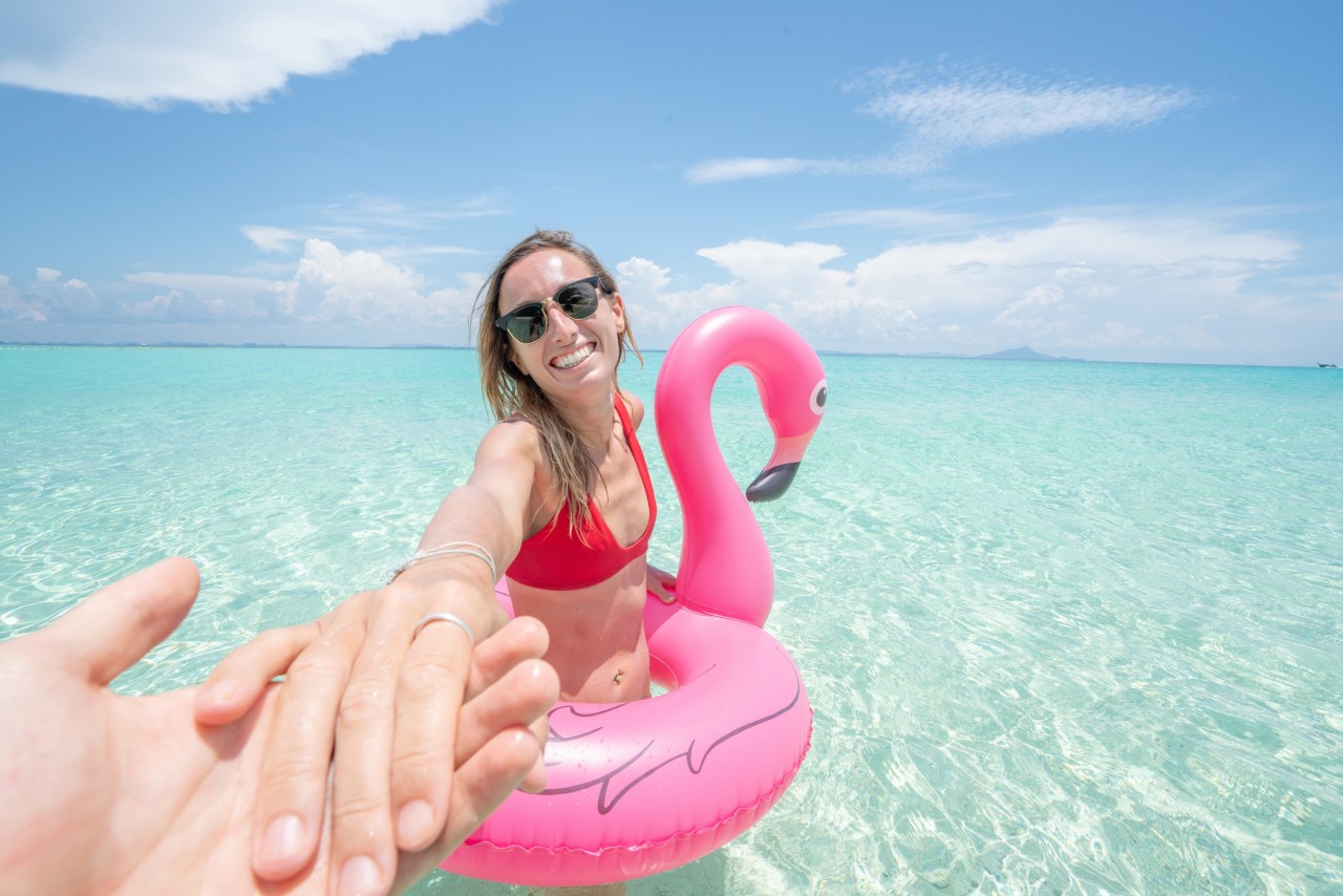 Follow me concept woman leading boyfriend on idyllic beach with inflatable flamingo playing in pristine clear water in the Islands of Thailand. People travel luxury fun and cool attitude concept