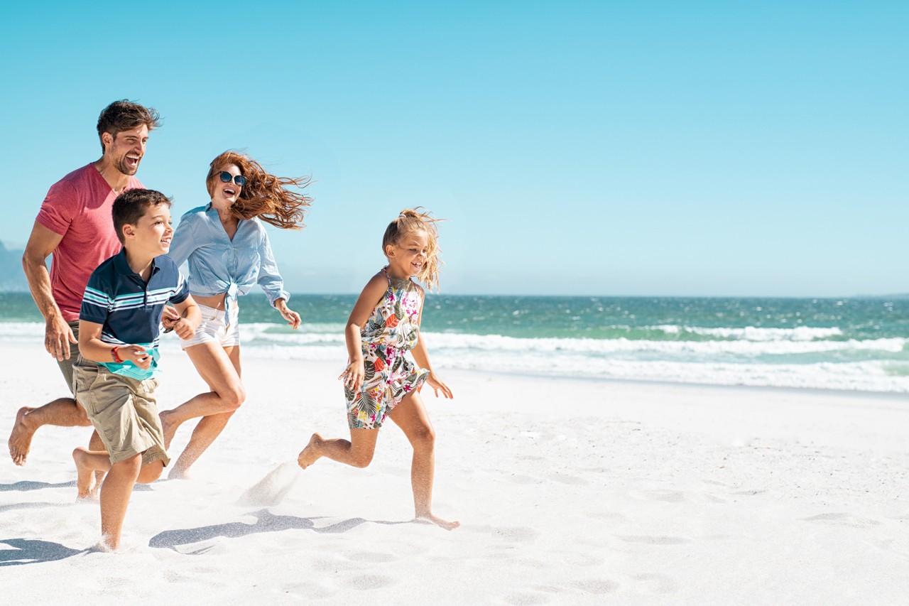 Cheerful young family running on the beach with copy space. Happy mother and smiling father with two children, son and daughter, having fun during summer holiday. Playful casual family enjoying playing at beach during vacaton.