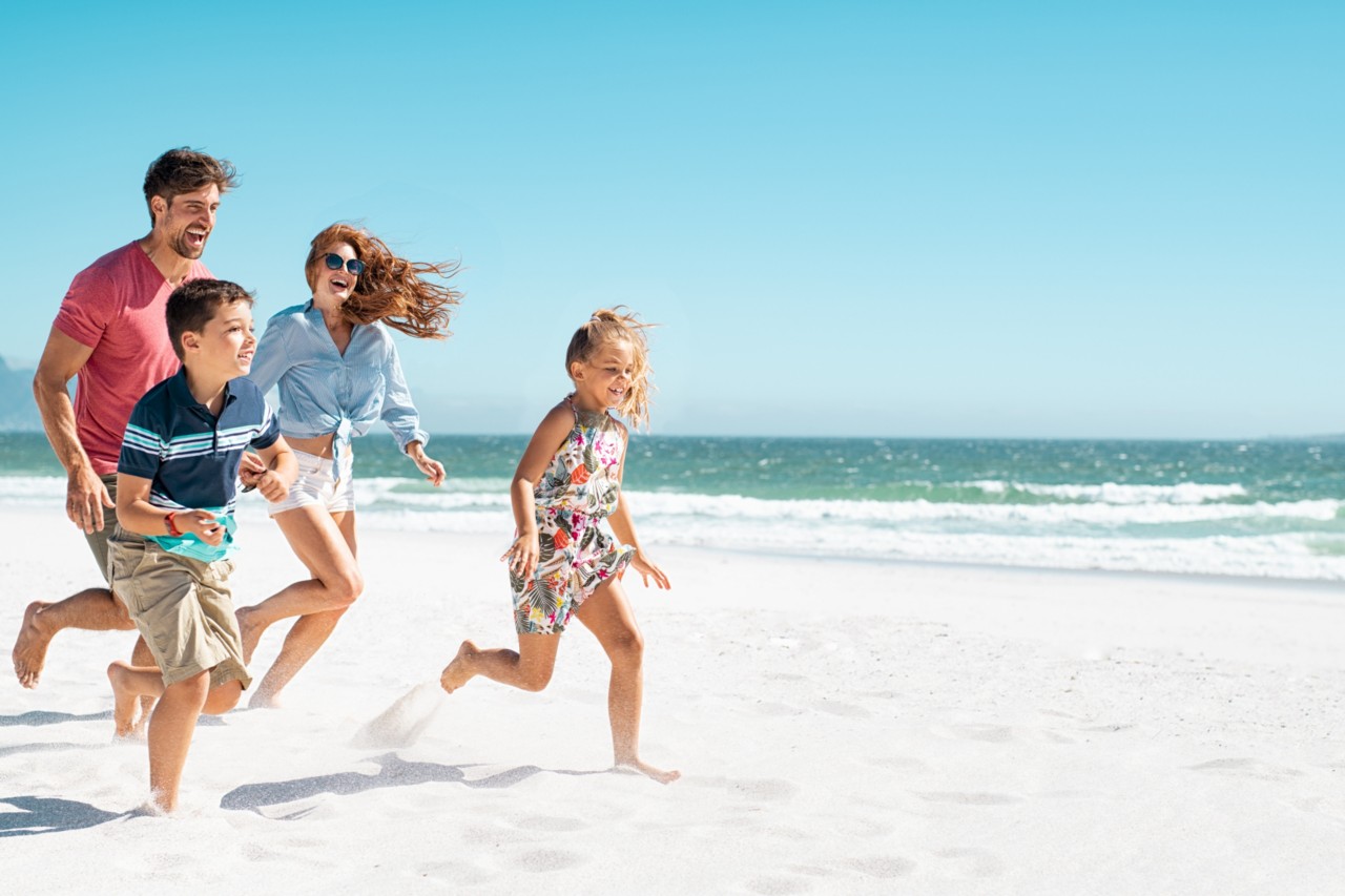 Cheerful young family running on the beach with copy space. Happy mother and smiling father with two children, son and daughter, having fun during summer holiday. Playful casual family enjoying playing at beach during vacaton.