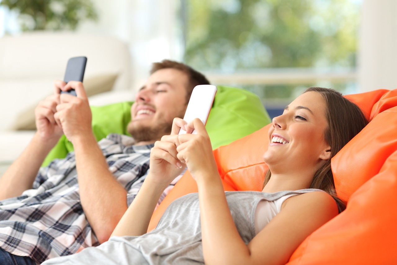 Couple using their smart phones lying on a green and orange poufs in the living room at home