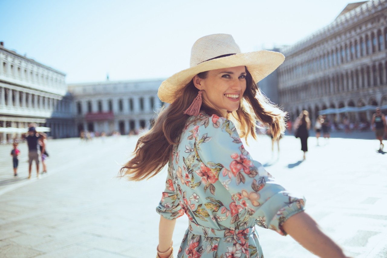 smiling trendy woman in floral dress with hat having excursion at San Marco square in Venice, Italy.