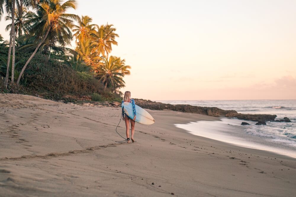 Surfen in der Karibik und Mittelamerika: Frau mit Surfbrett am Tropenstrand.
